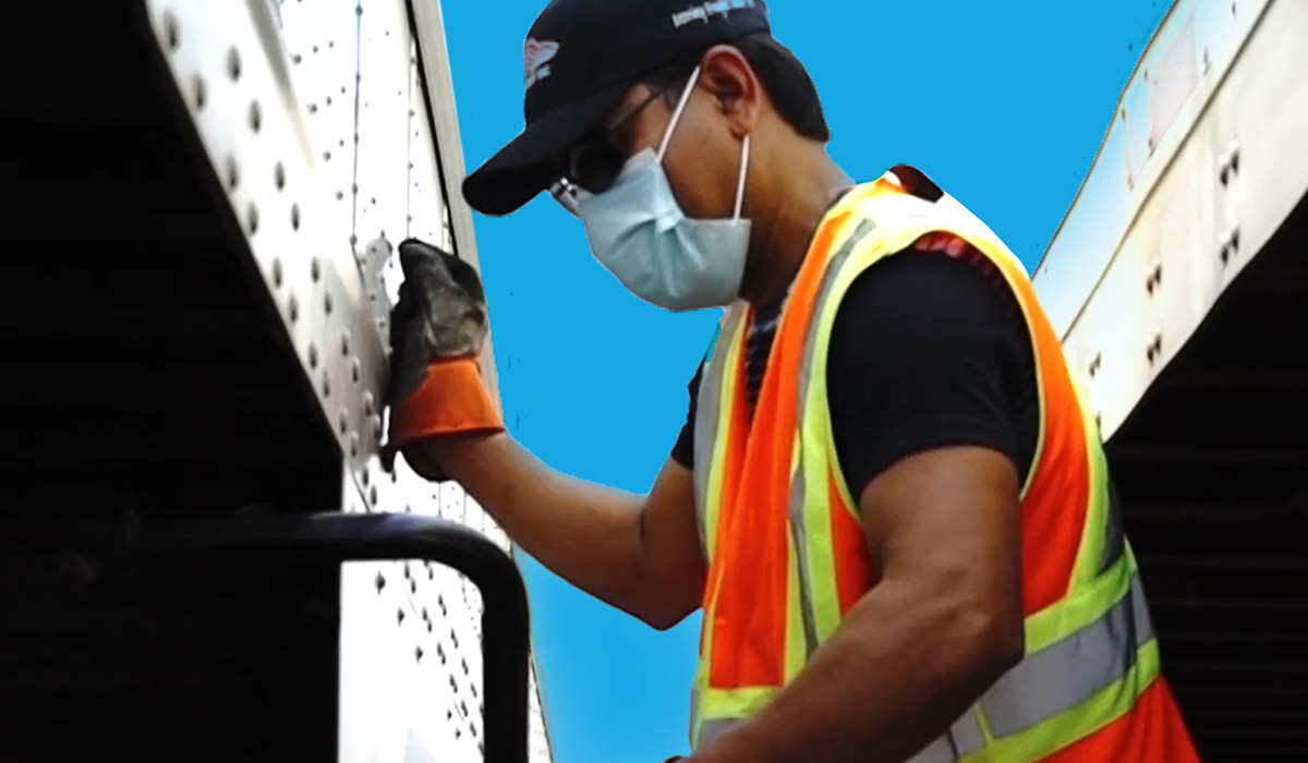 A KJS Transport crew member in a high-visibility vest and branded cap working on the side panel of a trailer