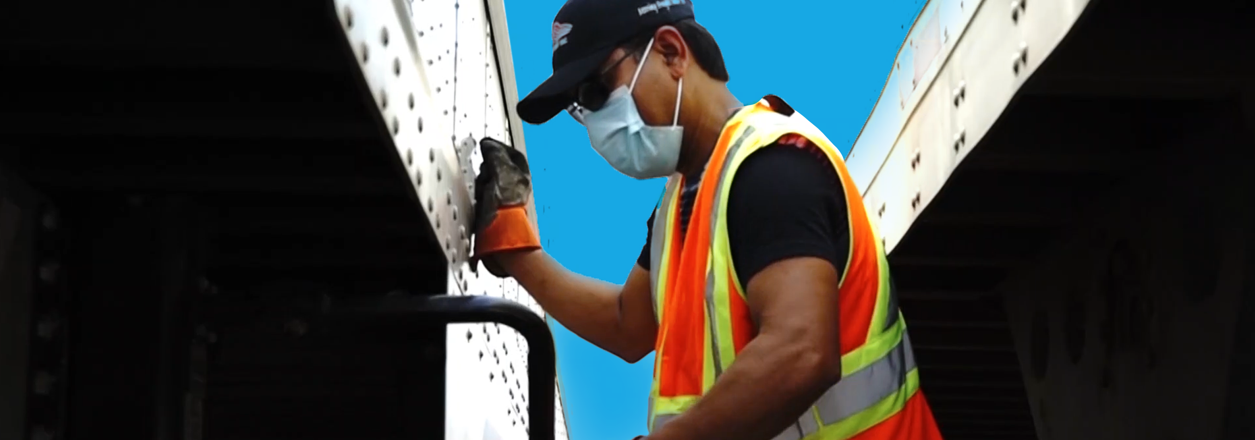 A KJS Transport crew member in a high-visibility vest and branded cap working on the side panel of a trailer