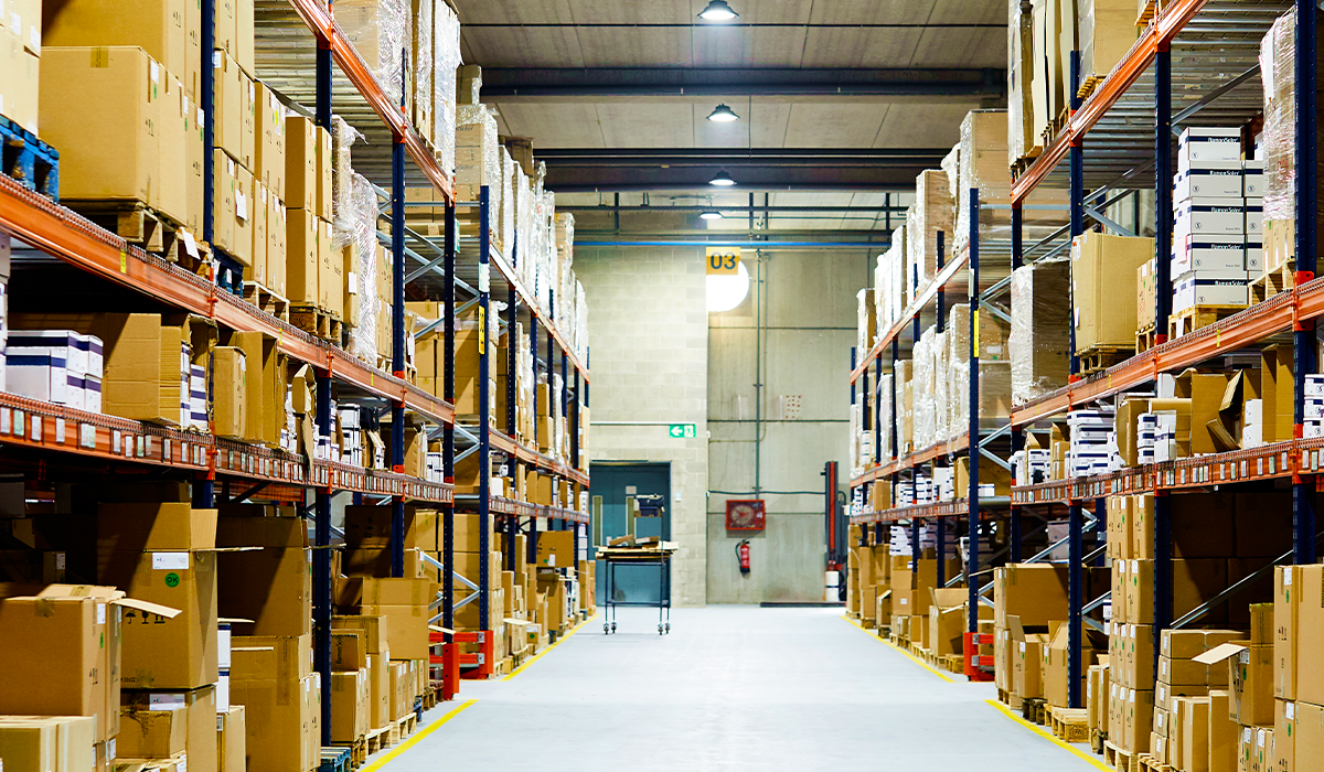 A wide aisle inside the KJS Transport distribution warehouse, lined floor to ceiling with palletised cartons on orange and blue racking