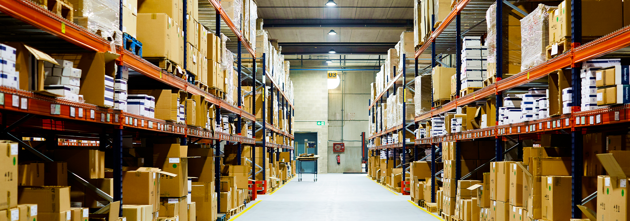 A wide aisle inside the KJS Transport distribution warehouse, lined floor to ceiling with palletised cartons on orange and blue racking